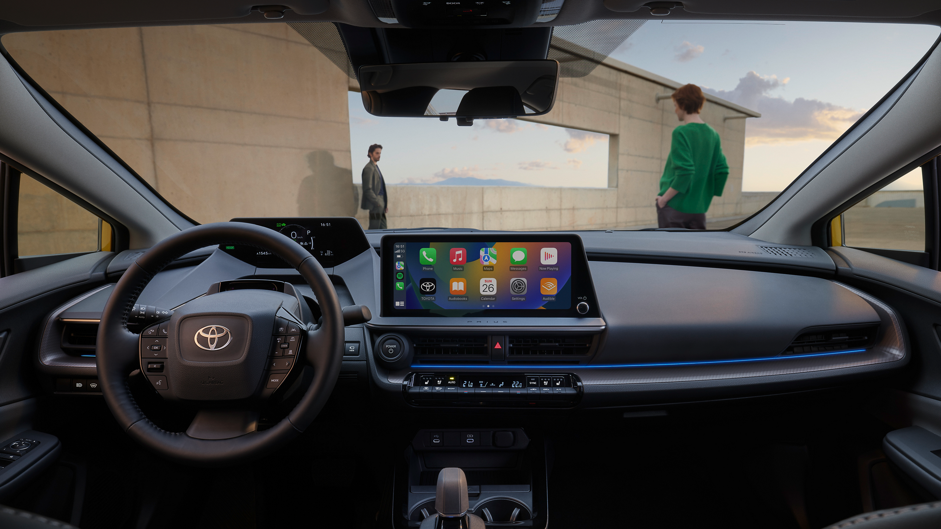 Blick auf das Cockpit des Prius mit dem Display und den Icons verschiedener Apps im Zentrum, durch die Frontscheibe sieht man eine Frau und einen Mann