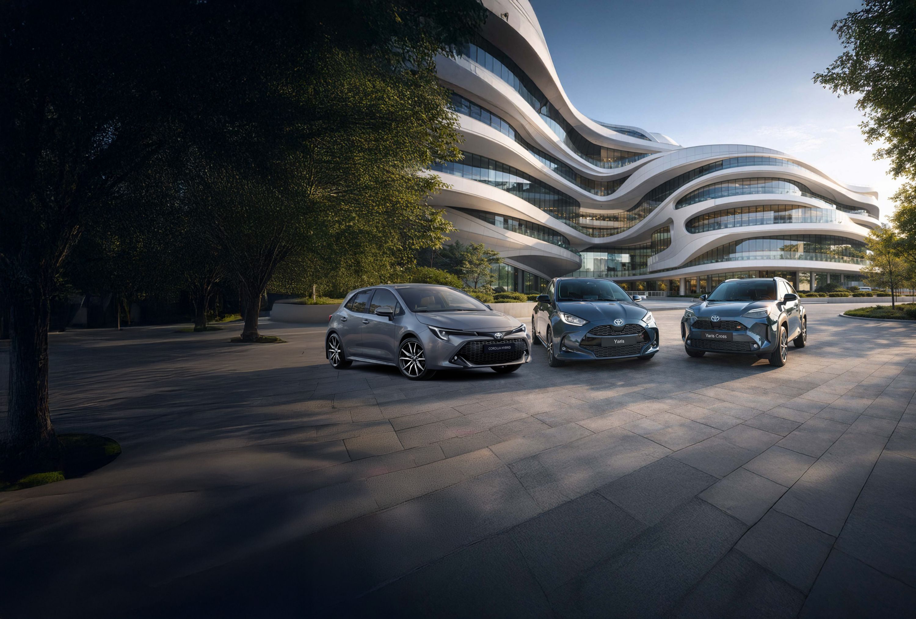 Ein Toyota Yaris und ein Toyota Yaris Cross stehen nebeneinander auf einem gepflasterten Platz vor modernen, terrassenförmig angeordneten Wohngebäuden bei Abendlicht.