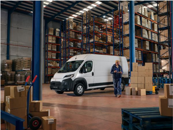 Großes Lagerhaus mit gefüllten Regalen und einem Arbeiter, der ein Klemmbrett hält und einem weißen Toyota Proace Max im Hintergrund.