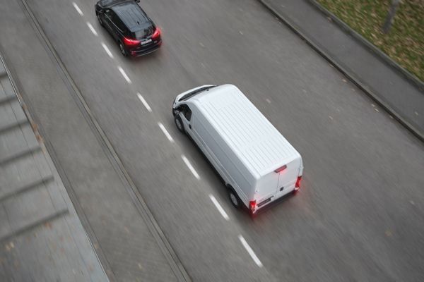 Vogelperspektive auf einen weißen Proace Max, der mit leuchtenden roten Rückleuchten hinter einem schwarzen Auto eine Straße entlangfährt.