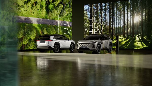 Scena in studio: un Toyota RAV4 PHEV argento in carica, un RAV4 Hybrid bianco parcheggiato accanto. Su due grandi schermi sullo sfondo vengono mostrati paesaggi forestali.
