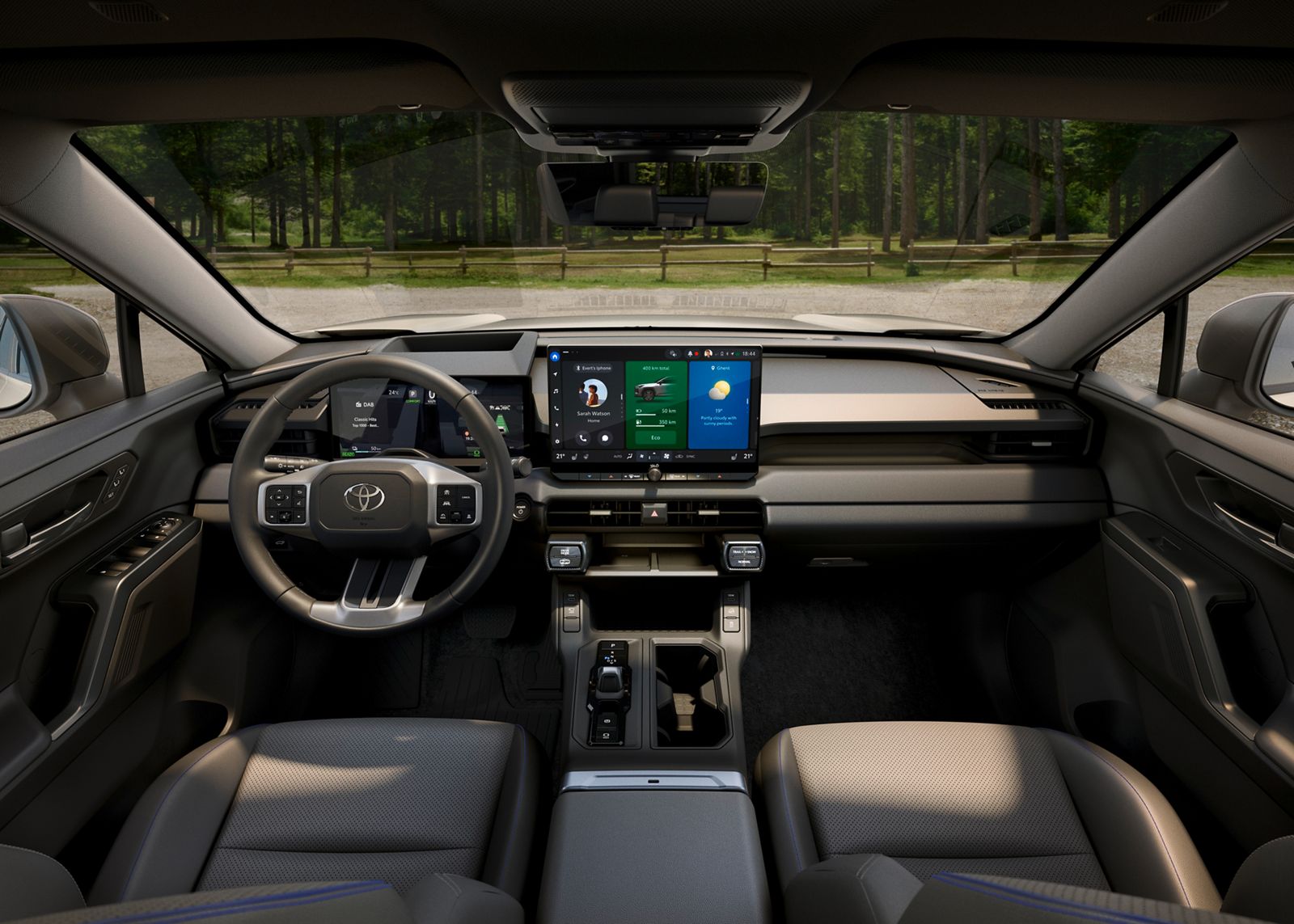 Vordersitze und Cockpit des RAV4 mit Multimedia-Display, Blick durch die Frontscheibe auf einen Schotterparkplatz, Zaun und Wald