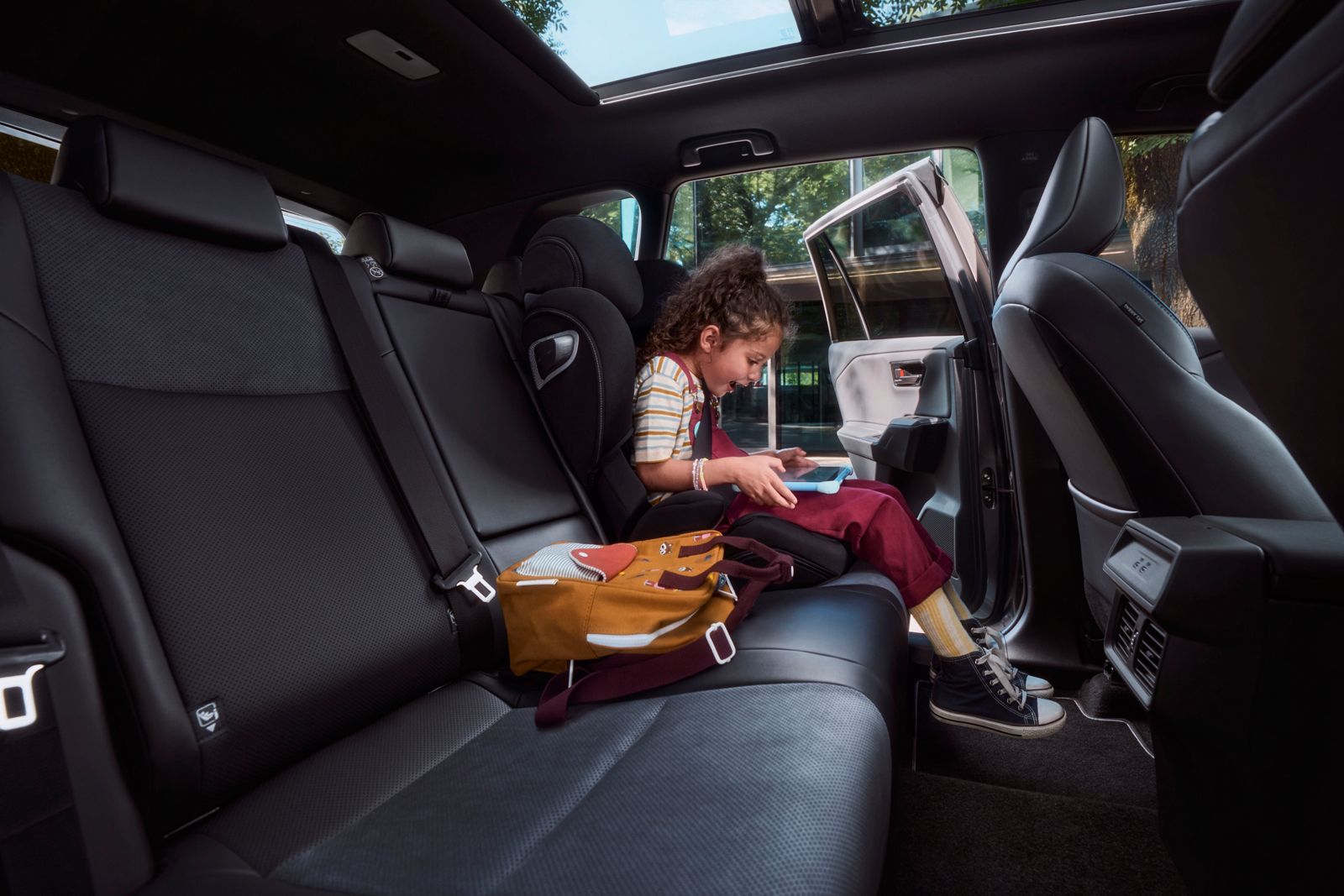 Rückbank des RAV4: Ein Mädchen sitzt im Kindersitz und schaut auf ihr Tablet, linke Tür geöffnet, Glasfassade im Hintergrund