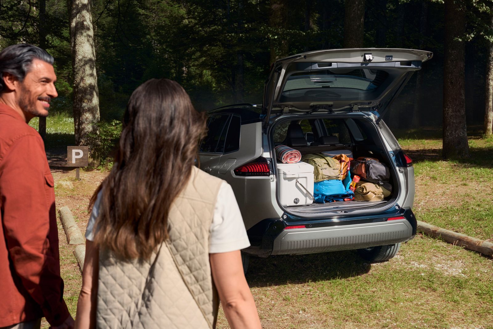 Geöffneter Kofferraum eines RAV4 mit Gepäck und Kühlbox, zwei Personen stehen daneben auf einem Waldparkplatz zwischen Bäumen