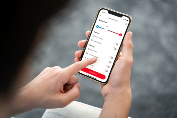 A person holds a smartphone displaying the MyToyota App. Their left hand taps the screen to pre-set their car’s climate control remotely.