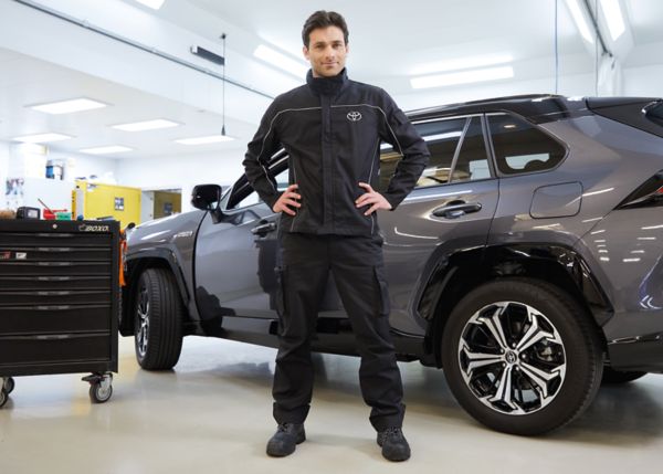 Ein Service Techniker im schwarzen Overall mit weißem Toyota-Logo auf der Brust, steht in einer Werkstatt vor einem grauen Toyota RAV4.
