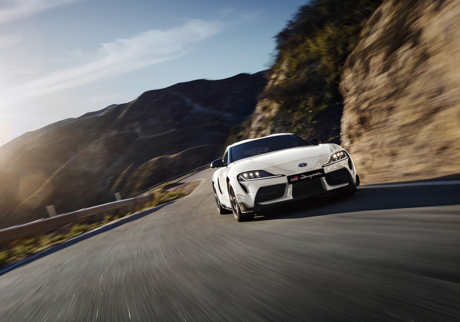 Ein weißer GR Supra von vorn dynamisch fotografiert während der Fahrt auf einer kurvigen Straße im Gebirge, im Hintergrund karg bewachsene Berge.