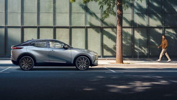 Silver Toyota C-HR+ electric SUV parked on an urban street. A man admires its design while walking by.