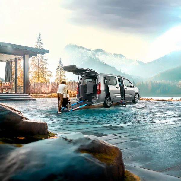 Umgebauter Proace City Verso steht vor einer Bergkulisse. Eine Person hilft einem Rollstuhlfahrer über eine Rampe in das Auto zu fahren.