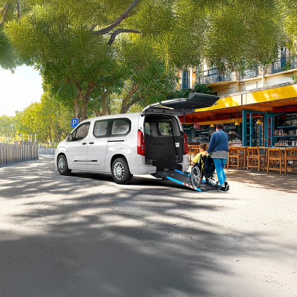 Person hilft einem Rollstuhlfahrer über eine Rampe in einen Proace City Verso hineinzufahren, der vor einem Restaurant geparkt ist.