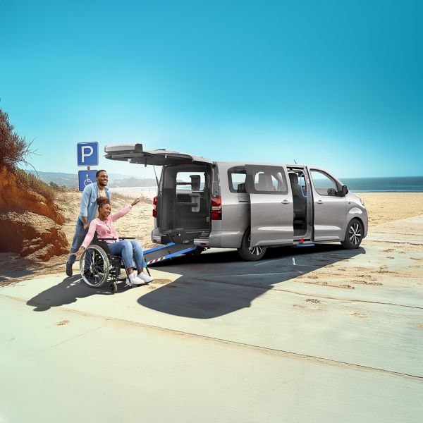Toyota Proace Verso steht an einem Strand mit ausgefahrener Laderampe, auf die eine Person im Rollstuhl mit der Hilfe einer weiteren Person fährt.