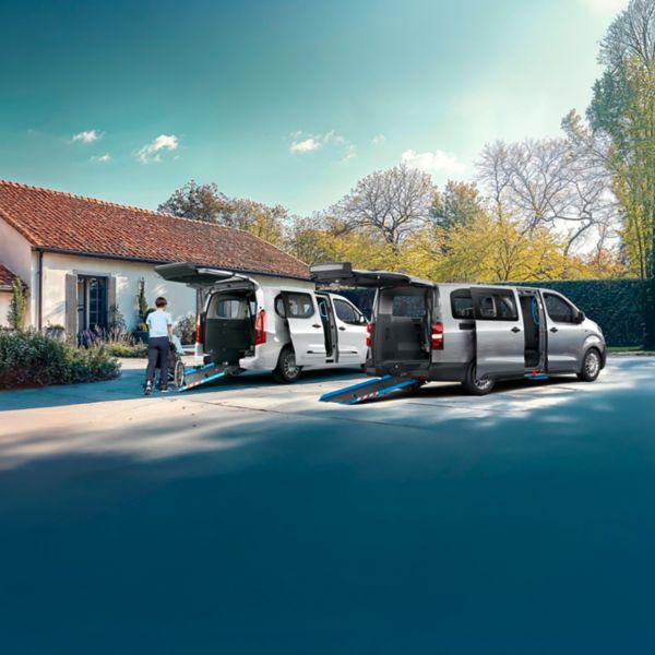 Zwei silberne Proace stehen auf einem Parkplatz vor einem Bungalow. Der Kofferraum ist geöffnet und eine Rampe ist ausgefahren, auf der ein Rollstuhlfahrer von einer zweiten Person hochgeschoben wird.