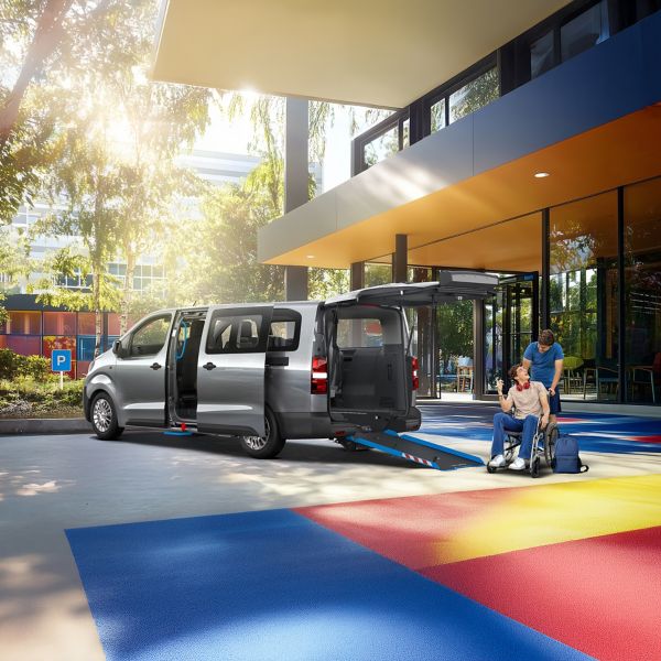 Proace Verso mit Laderampe steht auf einem bunten Parkplatz, zwei Personen unterhalten sich und eine hilft der anderen in den Proace Verso zu fahren.