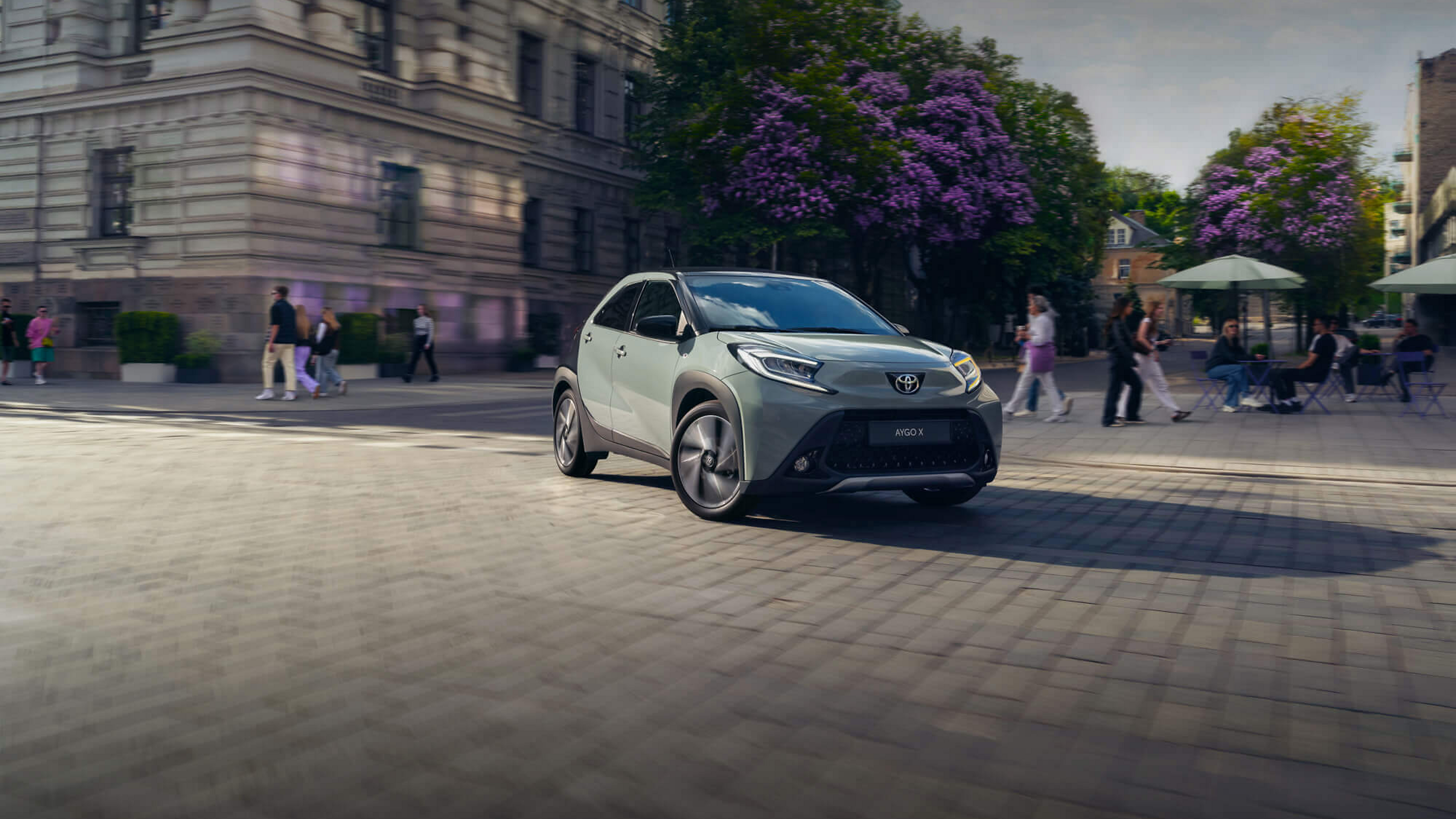 Mintfarbener Aygo X auf einem sonnigen Stadtplatz mit blühenden Bäumen und Menschen im Hintergrund.