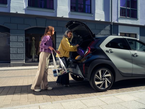 Zwei Frauen stehen auf einer gepflasterten Straße und laden ihre Taschen in den Kofferraum eines schwarzen Autos, das als Firmenwagen auch für private Fahrten genutzt werden kann.