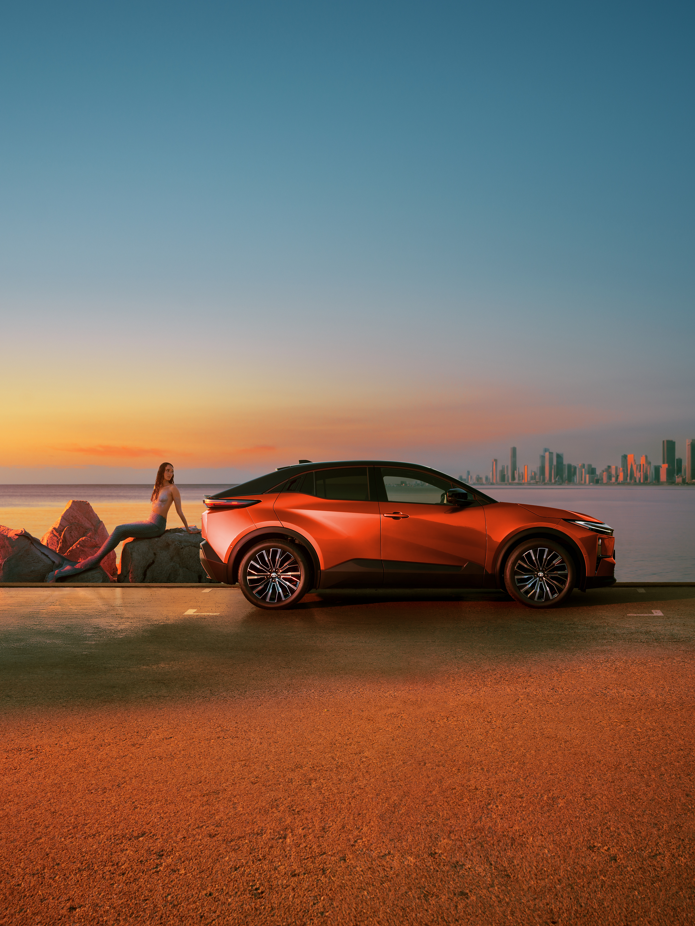 Orangefarbener C-HR+ steht bei Sonnenuntergang an einer Küstenstraße, im Hintergrund eine Skyline am Meer.