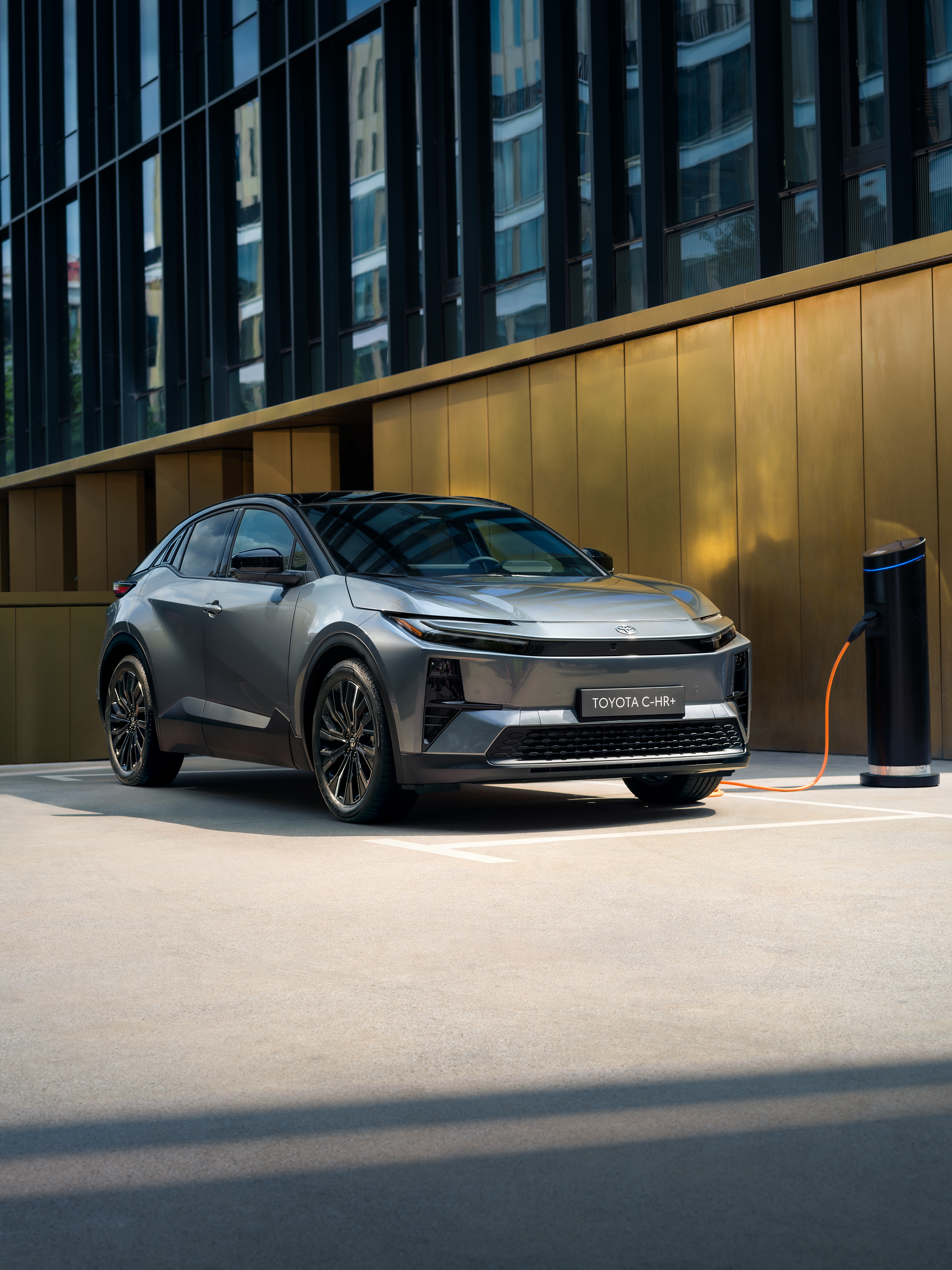 Silberner Toyota C-HR+ beim Laden an einer Ladesäule vor einem modernen Gebäude.
