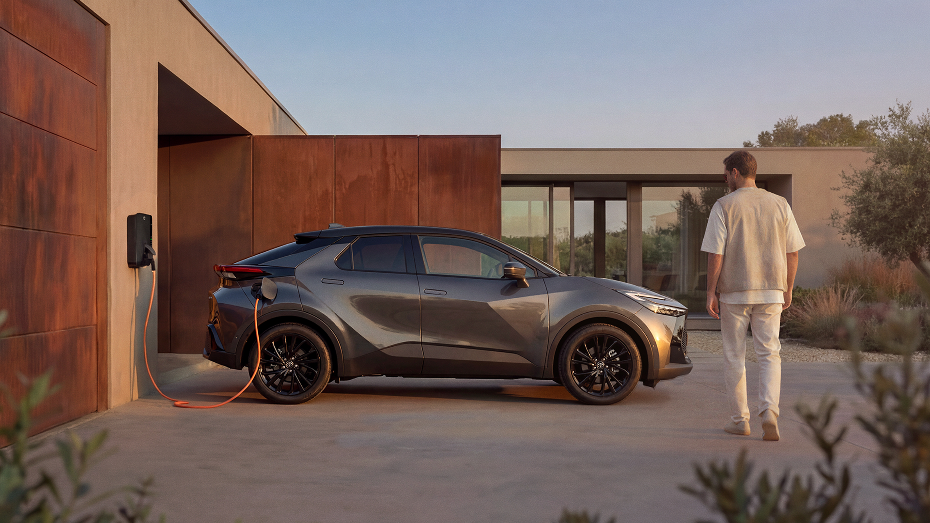 Ein Toyota C-HR lädt an einer Wallbox vor einem modernen Wohnhaus, während eine Person auf das Fahrzeug zugeht.