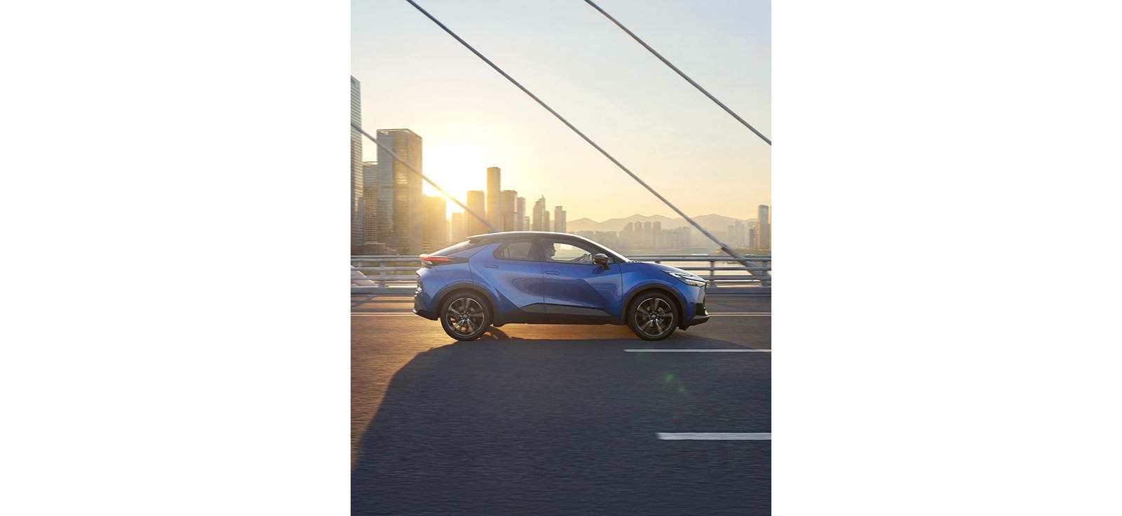 Ein blauer Toyota C-HR fährt bei Sonnenaufgang über eine Brücke vor moderner Stadtsilhouette.