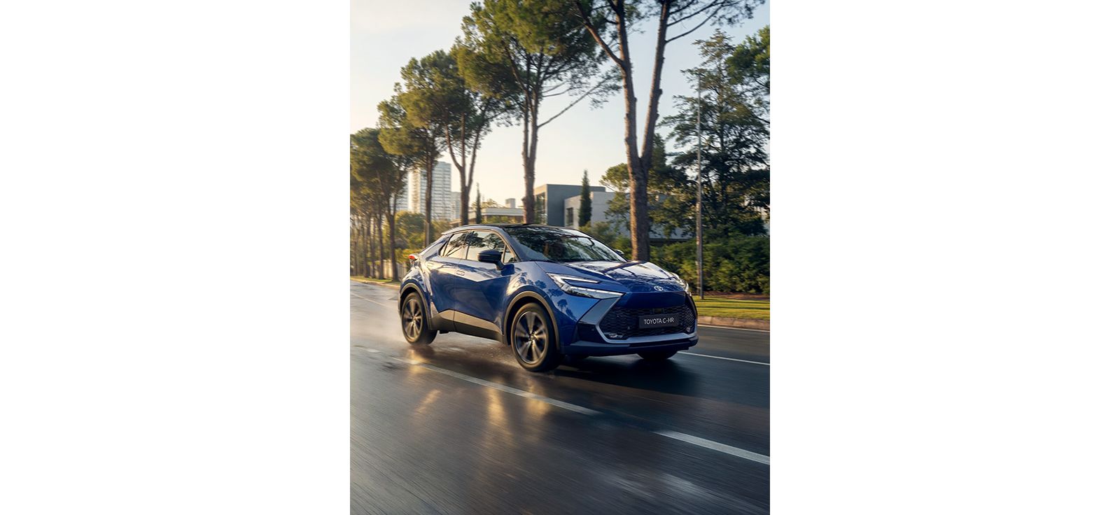 Ein blauer Toyota C-HR fährt auf einer nassen Straße durch eine grüne urbane Umgebung.