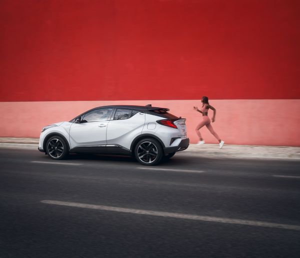 Ein weißer Kompakt-SUV parkt auf der Straße neben einer Frau in Sportkleidung, die vor einer leuchtend roten Wand joggt.
