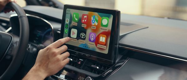 O persoana din interiorul unei Toyota interactioneaza cu ecranul tactil multimedia al masinii. Afisajul arata ecranul de pornire Apple CarPlay.