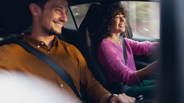Eine Frau am Steuer eines fahrenden Autos, ein Mann als Beifahrer, beide blicken lachend nach vorn 