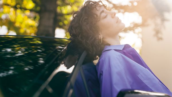 Eine Frau mit lockigem Haar lehnt sich mit geschlossenen Augen an ein Auto und genießt das Sonnenlicht in einer ruhigen Umgebung im Freien, die mit grünen Blättern gefüllt ist.