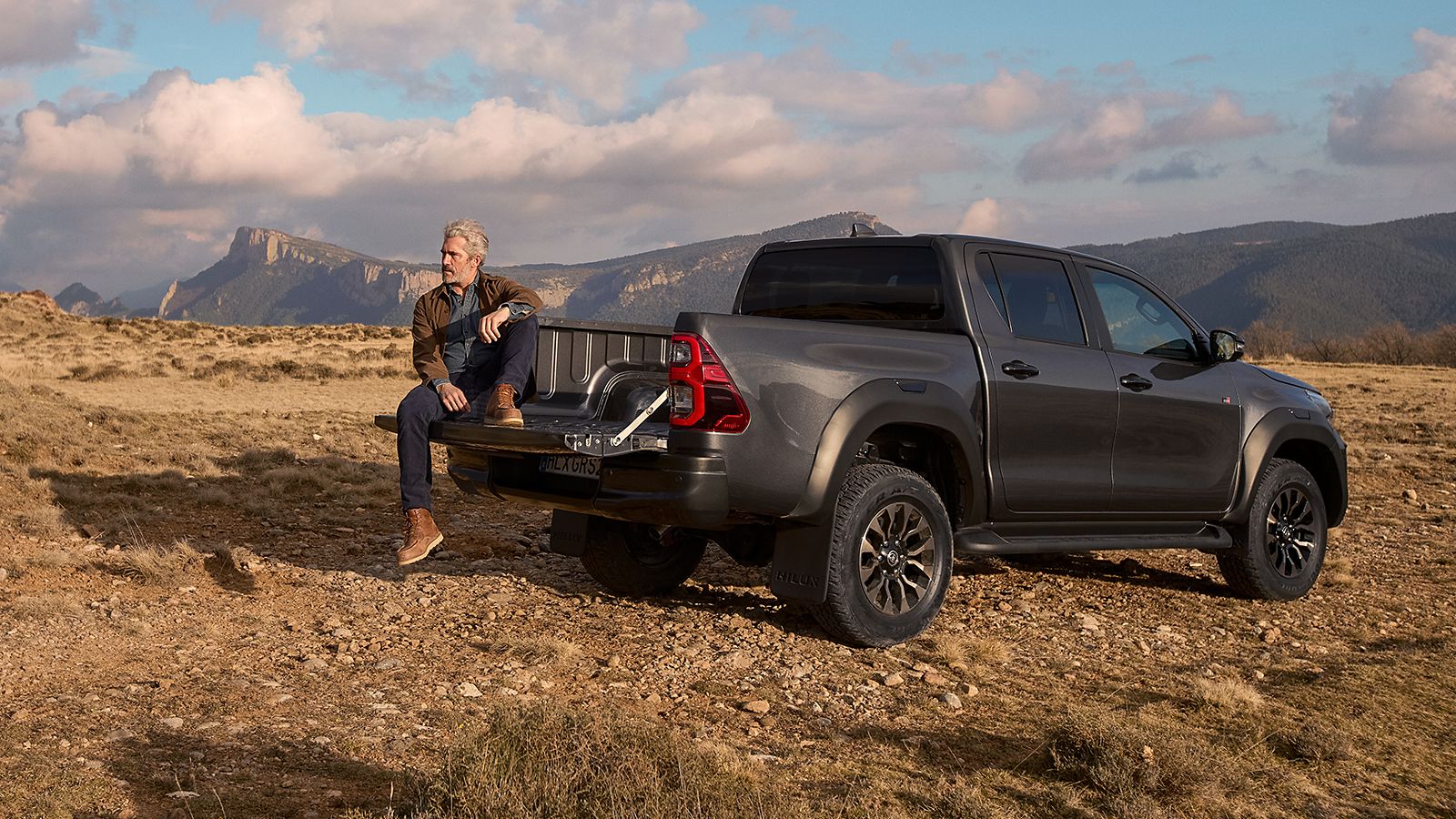 Eine Person sitzt hinten auf dem Toyota Hilux, im Hintergrund sind Berge und eine steinige Landschaft