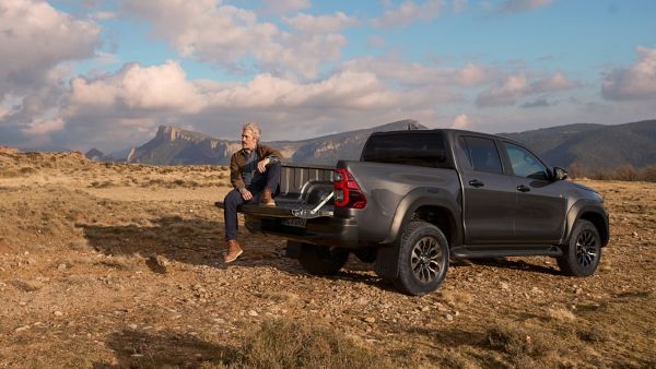 Eine Person sitzt hinten auf dem Toyota Hilux, im Hintergrund sind Berge und eine steinige Landschaft