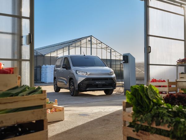 silbener Kleintransporter steht vor einem Gewächshaus an einer Ladesäule und lädt