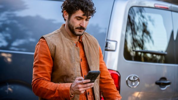 Eine Person mit einer Weste schaut auf ein Smartphone in der Hand. Dahinter sieht man das Heck eines Toyota Proaces