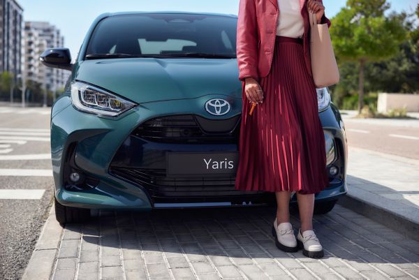 Eine Frau steht auf dem Gehsteig neben der Front ihres Firmenwagens und überlegt, wie sie die monatlichen Ausgaben für Versicherung und Steuer im Blick behalten kann.