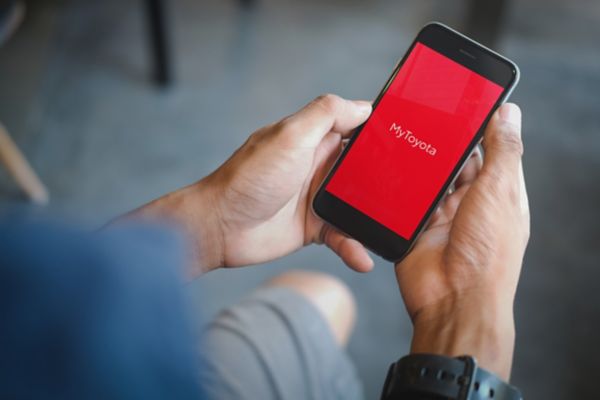 A hand holds aloft a smartphone showing the My Garage page of the TOYOTA MyT app. Available Connected Services are shown.