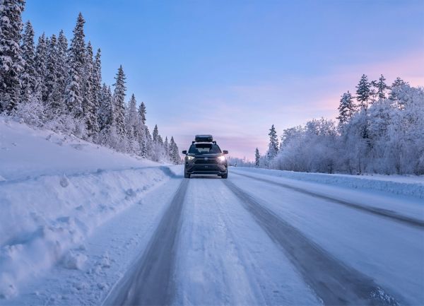 Toyota RAV4 Basis mit eingeschalteten Scheinwerfern fährt auf einer schneebedeckten Straße mit schneebedeckten Kiefern unter einem rosafarbenen Himmel.