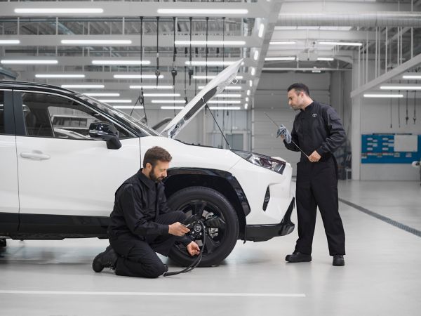 Zwei Mechaniker überprüfen parallel den Reifendruck und Ölstand eines weißen Toyota RAV4 im hellen Neonlicht einer modernen Werkstatt