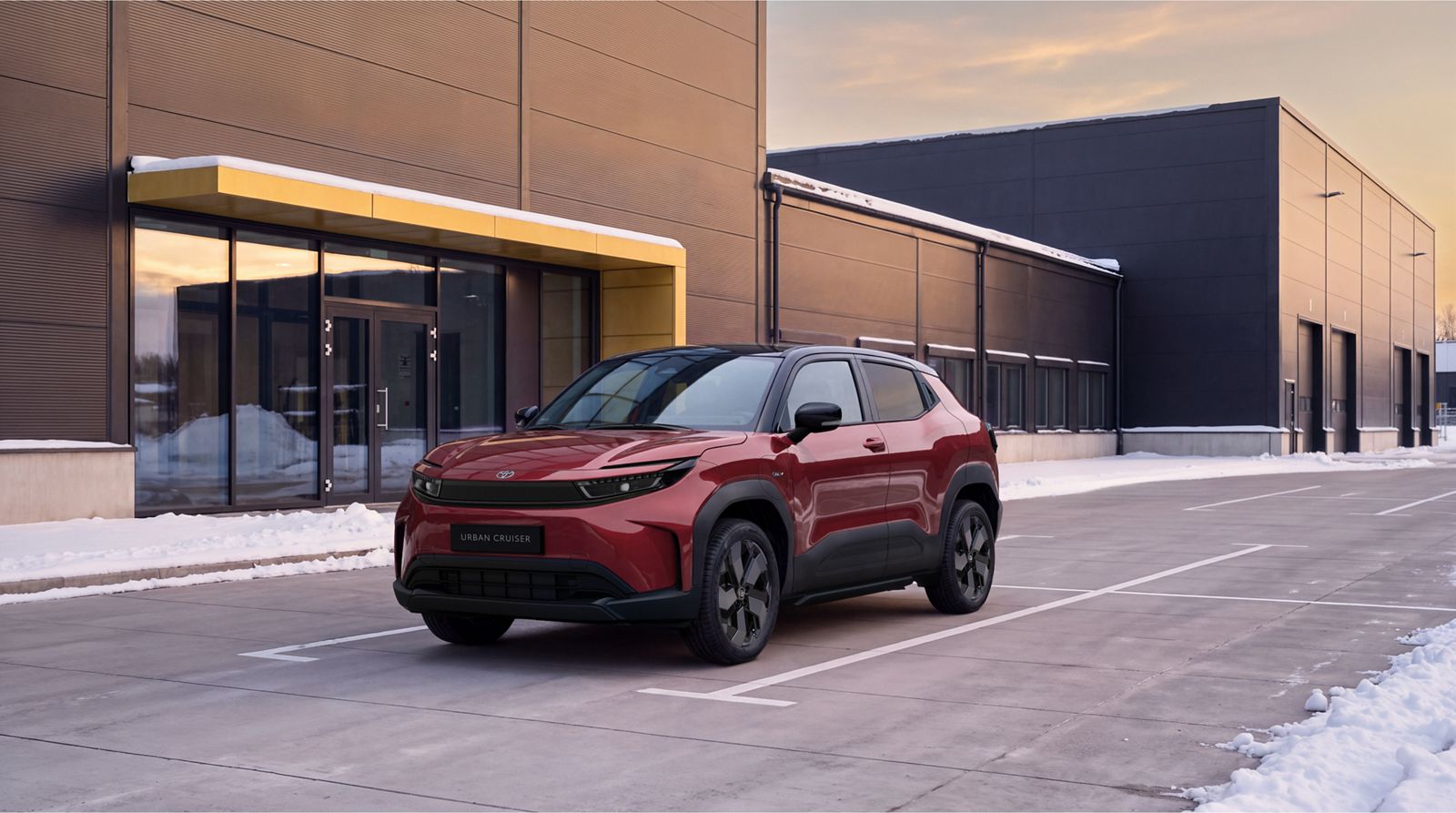 Roter Toyota Urban Cruiser steht auf einem Parkplatz vor einem modernen Gebäude mit großen Fenstern und gelbem Vordach bei winterlichen Bedingungen mit Schnee am Rand.
