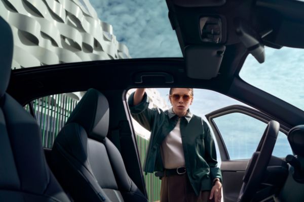 Una mujer elegantemente vestida apoya la mano en el techo de un Toyota. La puerta del coche está abierta, y los edificios son visibles a través de su a través de su gran techo de cristal.