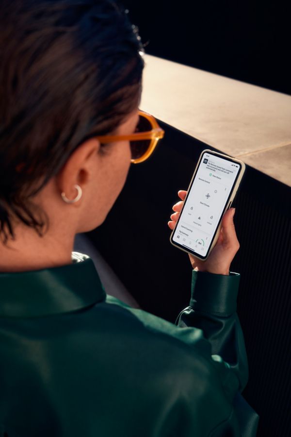 Mirando por encima del hombro de una mujer con chaqueta verde y gafas de sol. Sostiene un smartphone que muestra la página de inicio de la aplicación MyToyota.