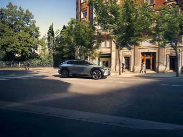 Ein moderner silberfarbener SUV fährt eine von Bäumen gesäumte Straße entlang, vorbei an einem historischen Gebäude mit Fußgängern in der Nähe, unter einem klaren Himmel.