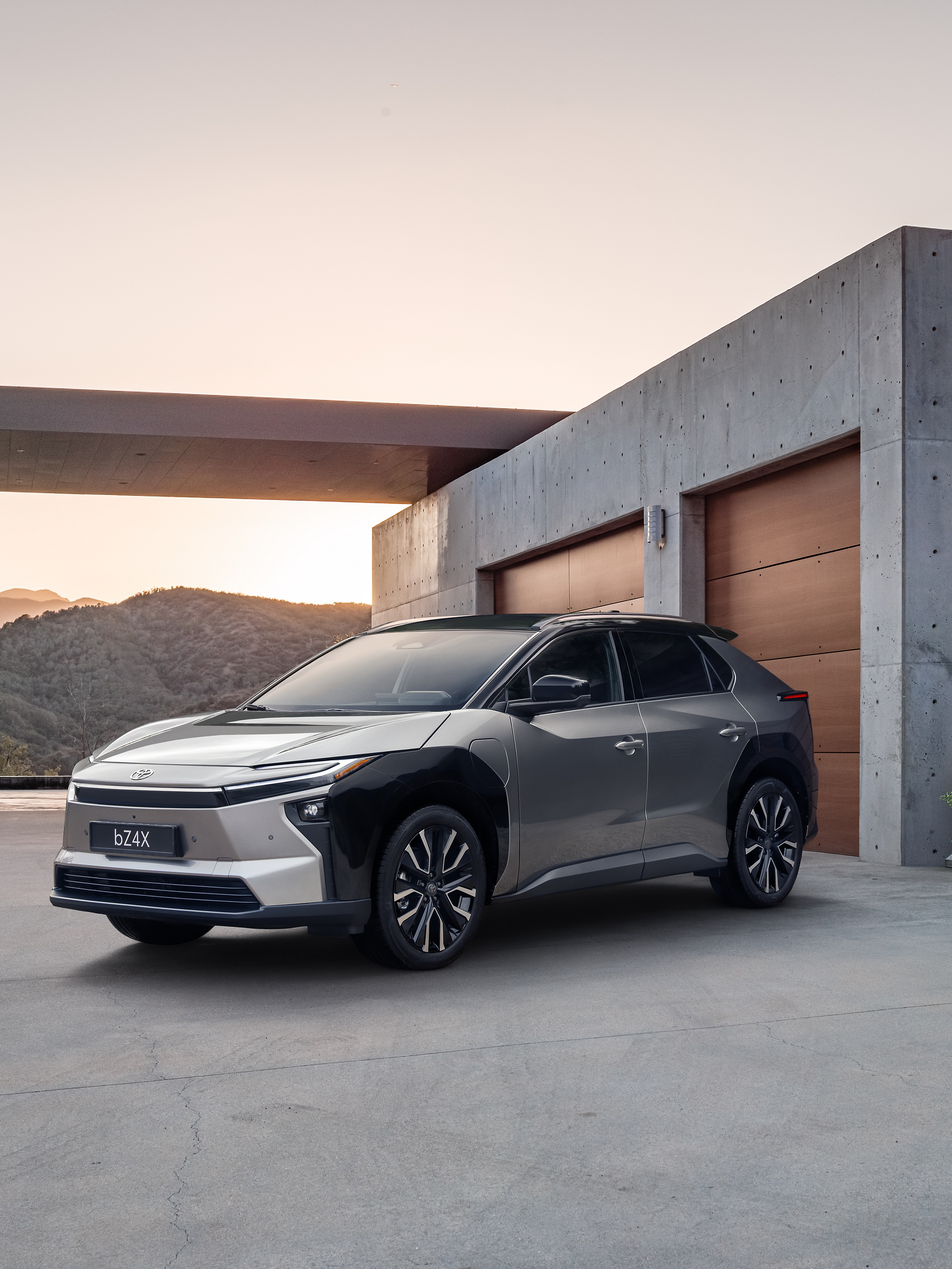 Grauer Toyota bZ4X Elektro-SUV steht vor einem modernen, minimalistischen Betonhaus mit Garage, im Hintergrund hügelige Landschaft bei warmem Abendlicht.