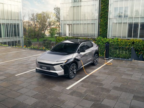 Ein silberner Toyota bZ4X steht auf einem Parkplatz vor einem modernem Bürogebäude und lädt an einer Ladesäule.