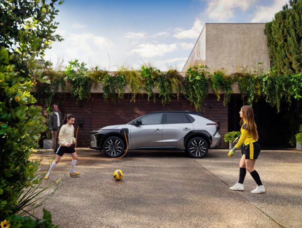 Ein grauer Toyota bZ4X  steht in der Einfahrt eines modernen Hauses und lädt über eine Wallbox, während Kinder im Vordergrund Fußball spielen.