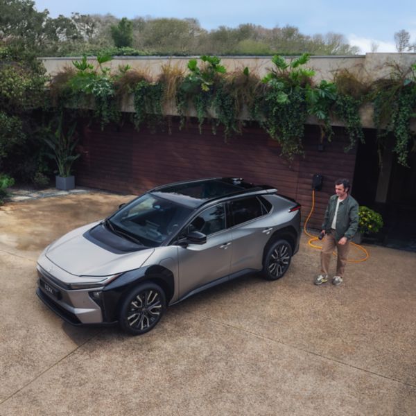 Silbernes E-SUV steht auf einer Hauseinfahrt und lädt an einer Wallbox. Eine Person läuft auf das Auto hinzu.