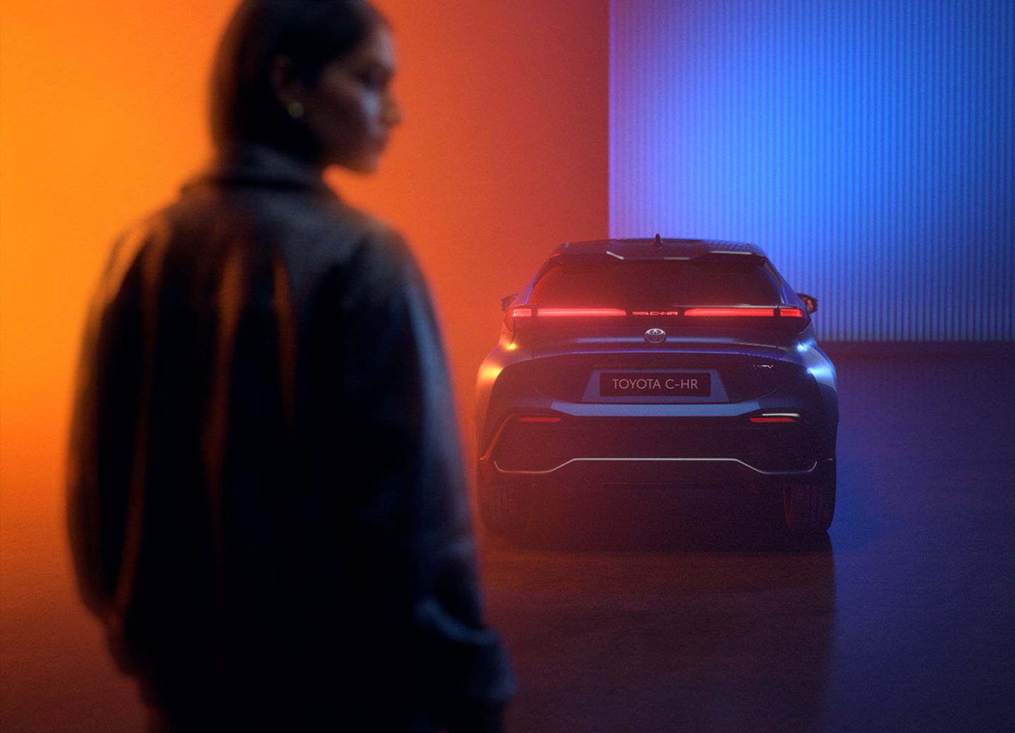 Rückansicht eines dunklen Toyota C-HR mit rot leuchtendem Rückleuchtenband und Kennzeichen „Toyota C-HR“ in einer Halle mit orange-blauer Beleuchtung, im Vordergrund unscharf eine Person von hinten.