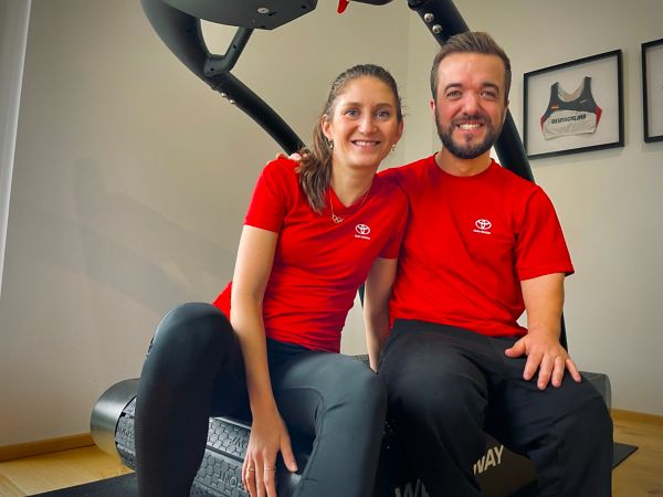 Zwei lächelnde Personen in passenden roten Toyota T-Shirts sitzen zusammen auf einem Fitnessgerät in einem hellen Raum.