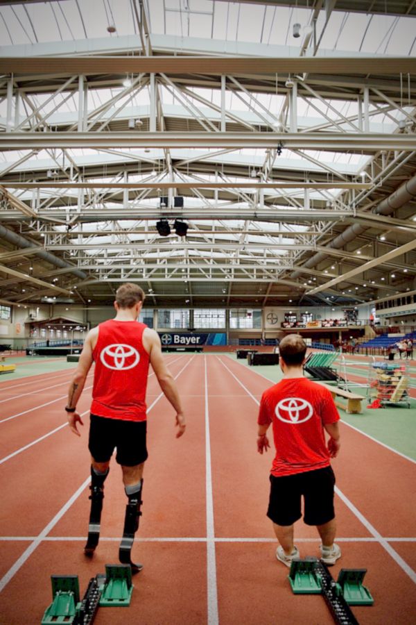 Zwei Athleten, einer mit Beinprothesen, in roten Shirts mit Toyota-Logo bereiten sich auf ein Rennen auf einer Indoor-Bahn vor.
