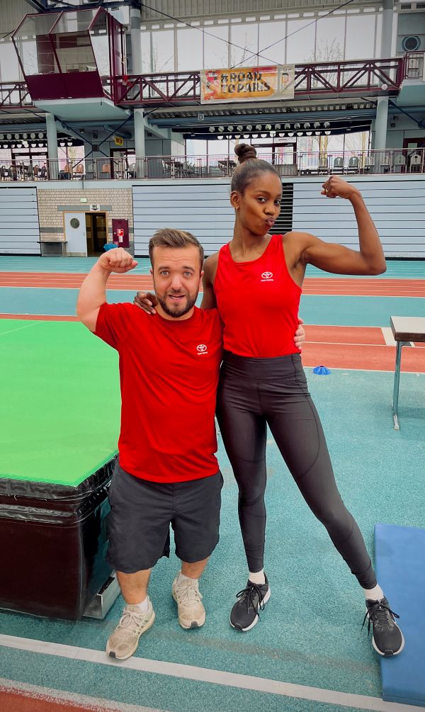 Zwei Athleten in roten Tanktops stehen in einer Sportanlage, im Hintergrund ist Sportausrüstung zu sehen.