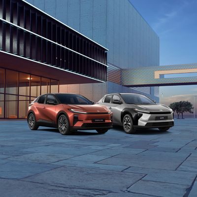 Drei Toyota-Fahrzeuge in Schwarz, Orange und Grau vor einem farbigen, grafischen Hintergrund, nebeneinander auf einer stilisierten Fläche präsentiert.