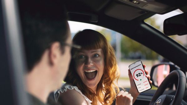 Junge Frau am Steuer eines Toyota zeigt ihrem Beifahrer begeistert lachend ihr Smartphone mit einem „Hybrid Coaching Score“ von 92 %.